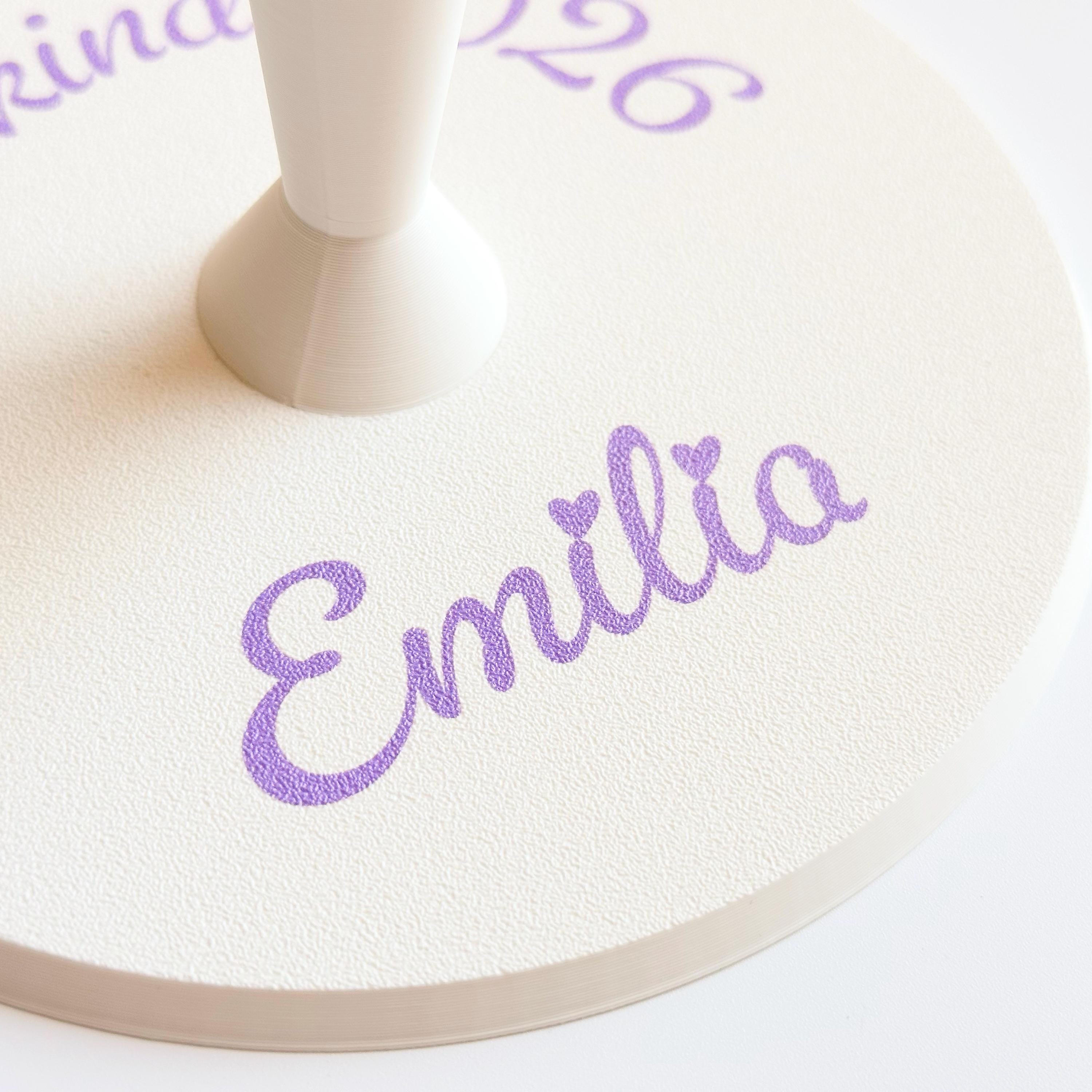 A close-up of the cream-colored round base shows the purple script text "Emilia" and the bottom of the conical stand.