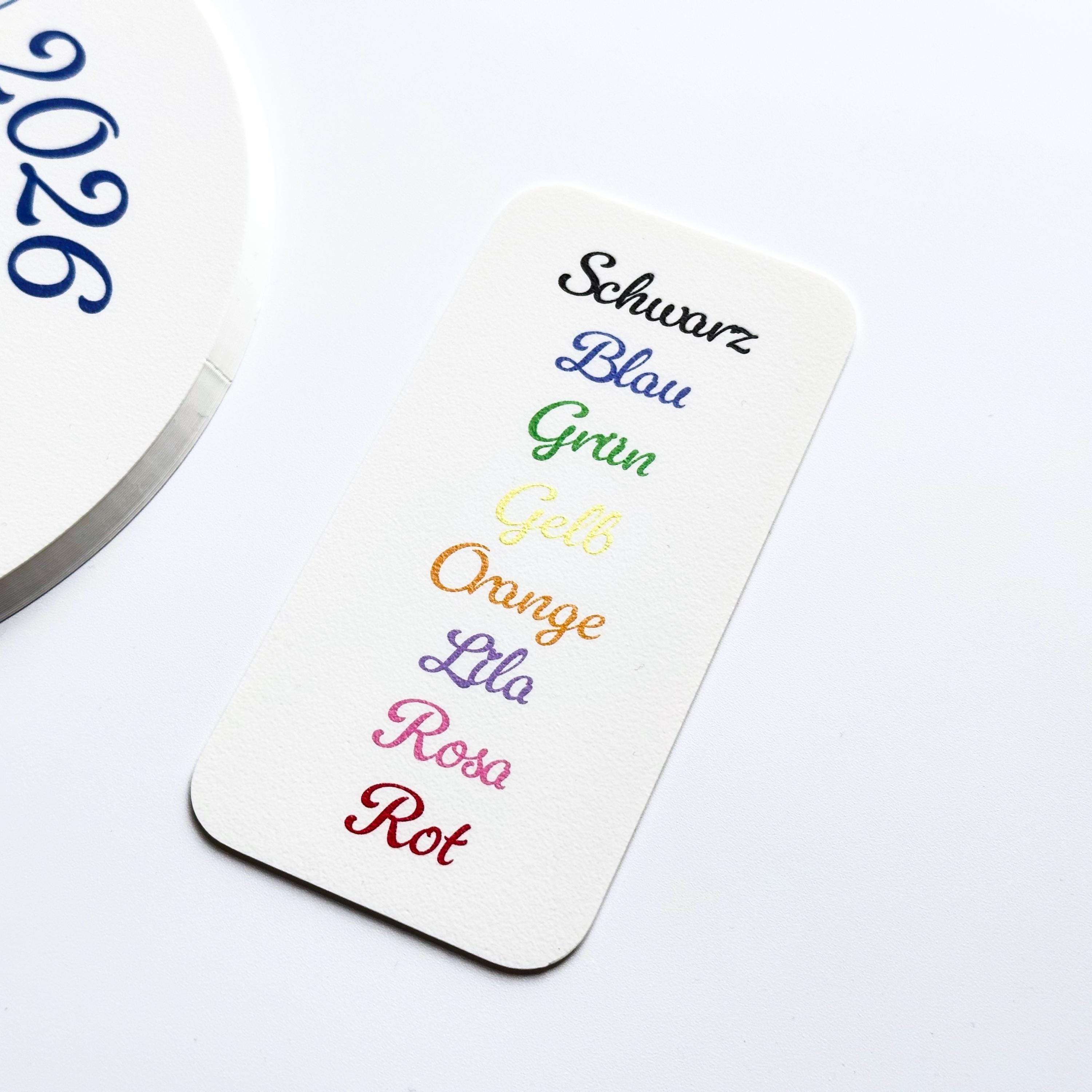 A white rectangular card displays a list of German color names, each printed in its respective color: Schwarz, Blau, Gruen, Gelb, Orange, Lila, Rosa, Rot.