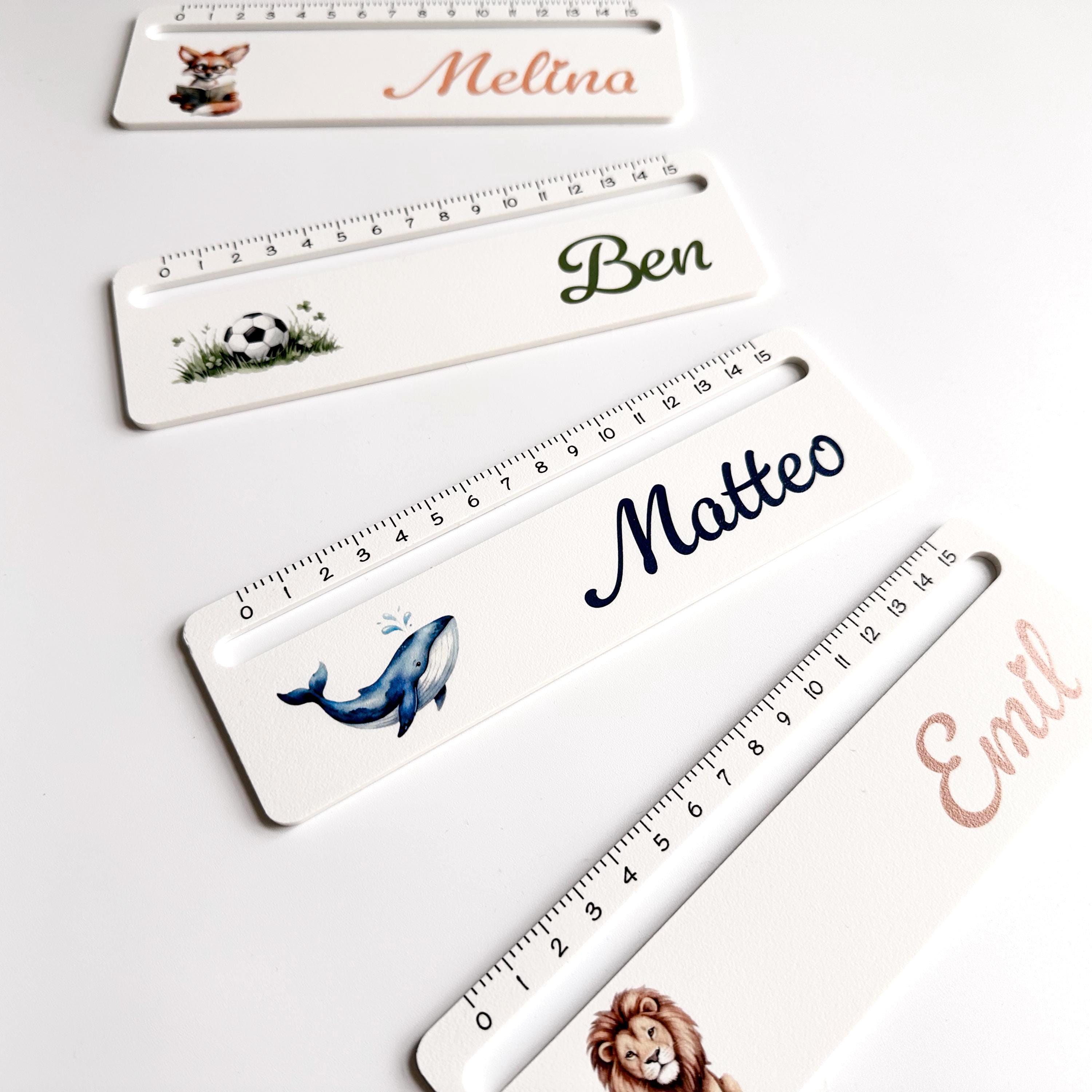 A close-up, angled view of four white rulers with black measurement markings, each personalized with a name and a different colorful illustration, on a light brown surface.