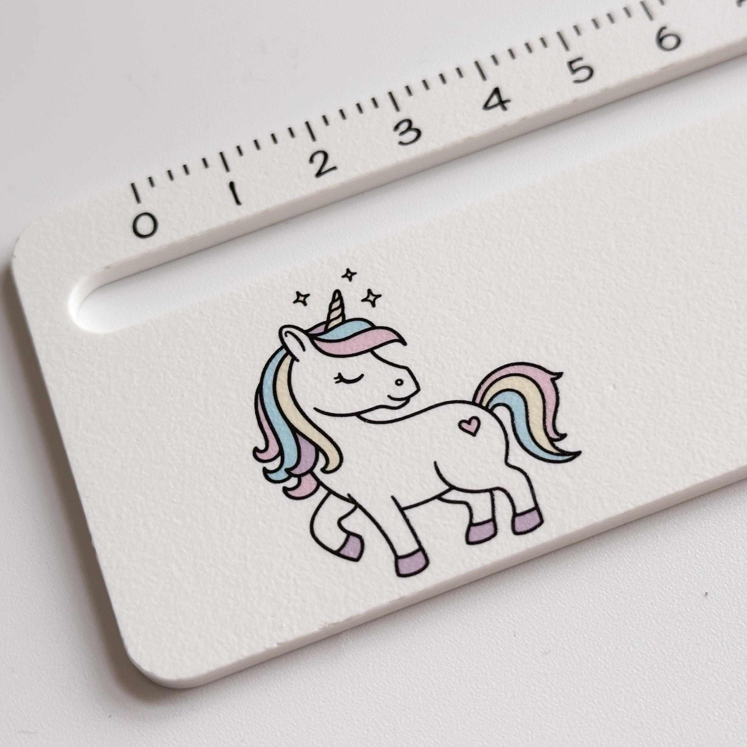 A close-up of a white ruler with black measurement markings, featuring a colorful unicorn illustration with a rainbow mane and tail, and small stars.