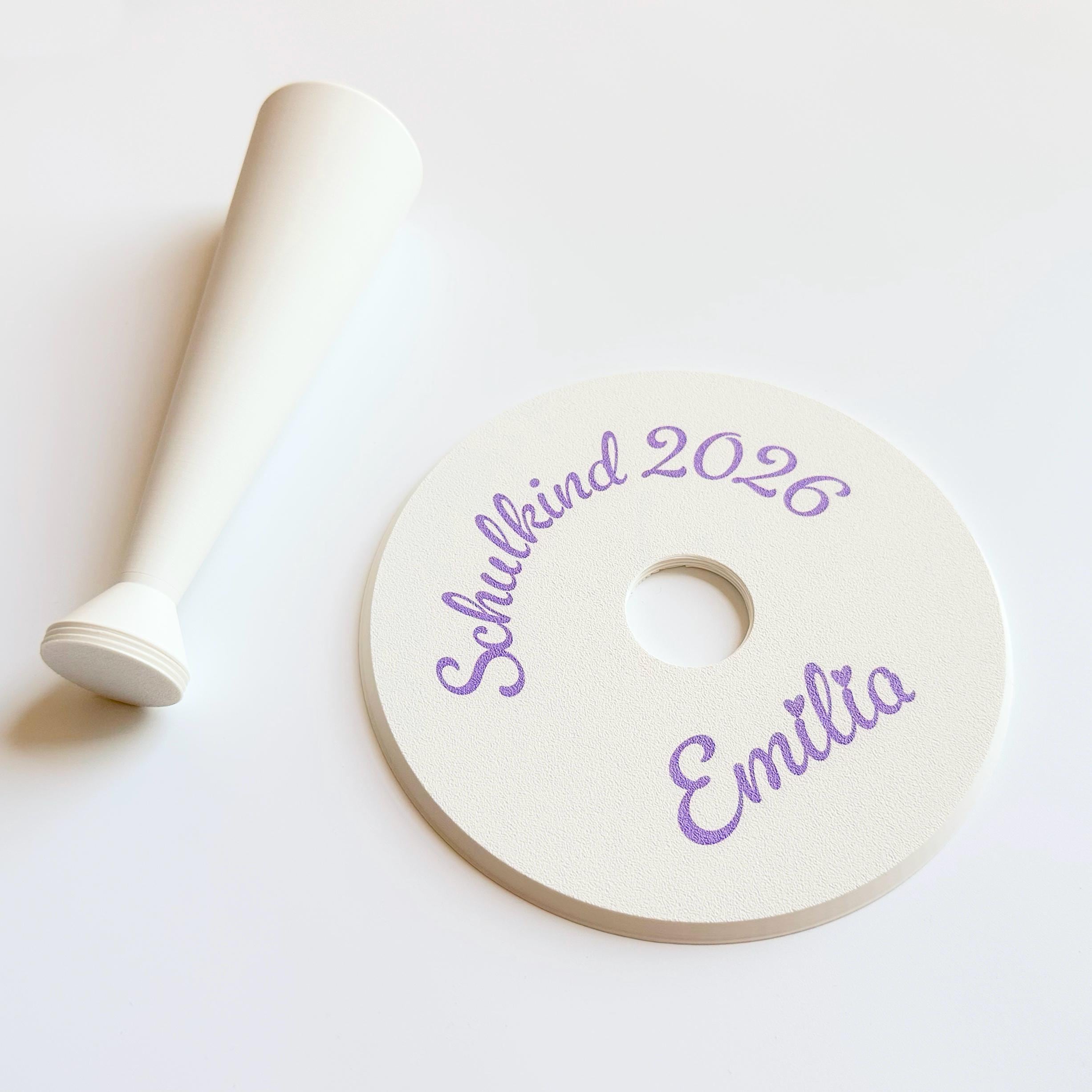 A cream-colored conical stand lies next to a cream-colored round base with a central hole, both with purple script text "Schulkind 2026" and "Emilia" on the base.