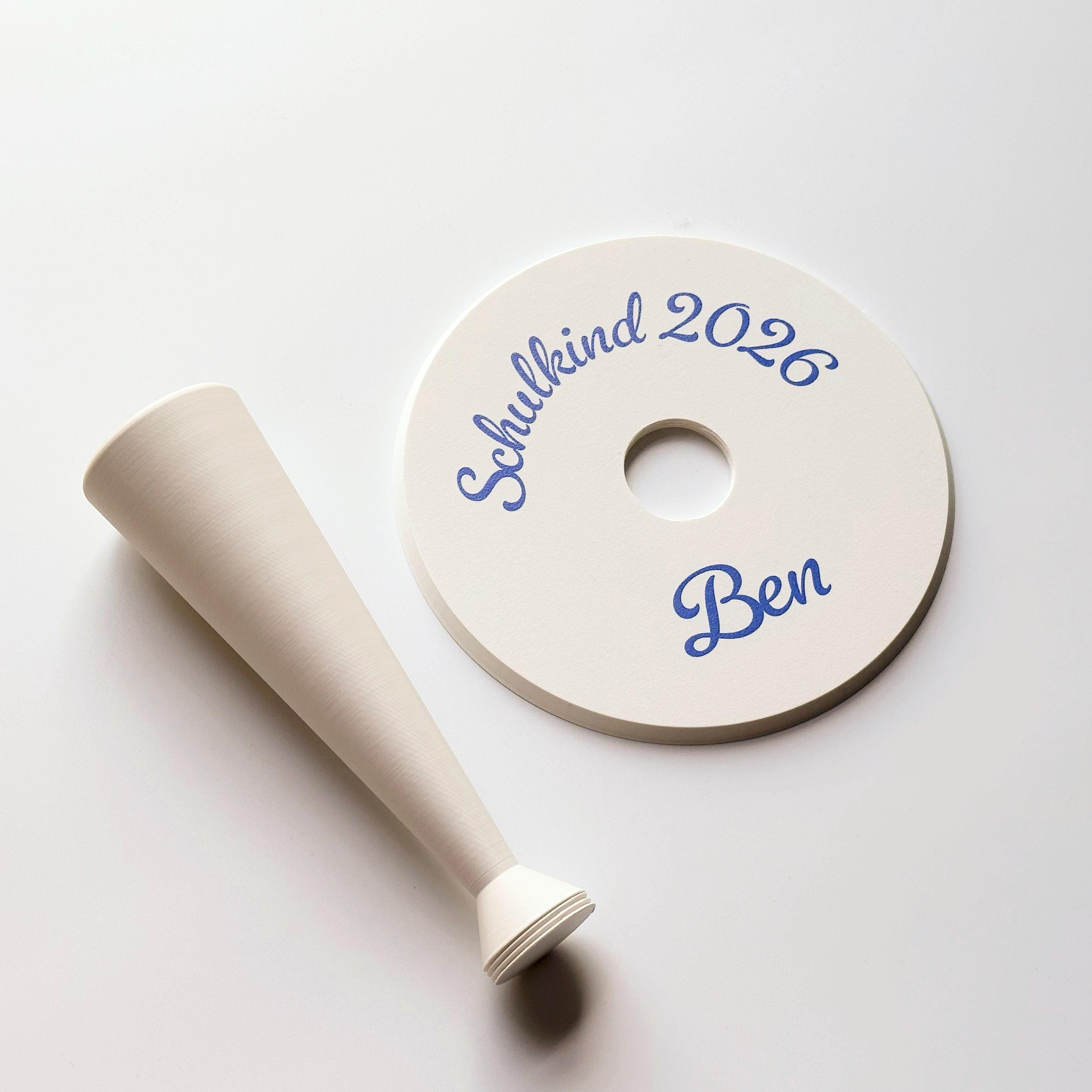 A cream-colored conical stand lies next to a cream-colored round base with a central hole, both with blue script text "Schulkind 2026" and "Ben" on the base.