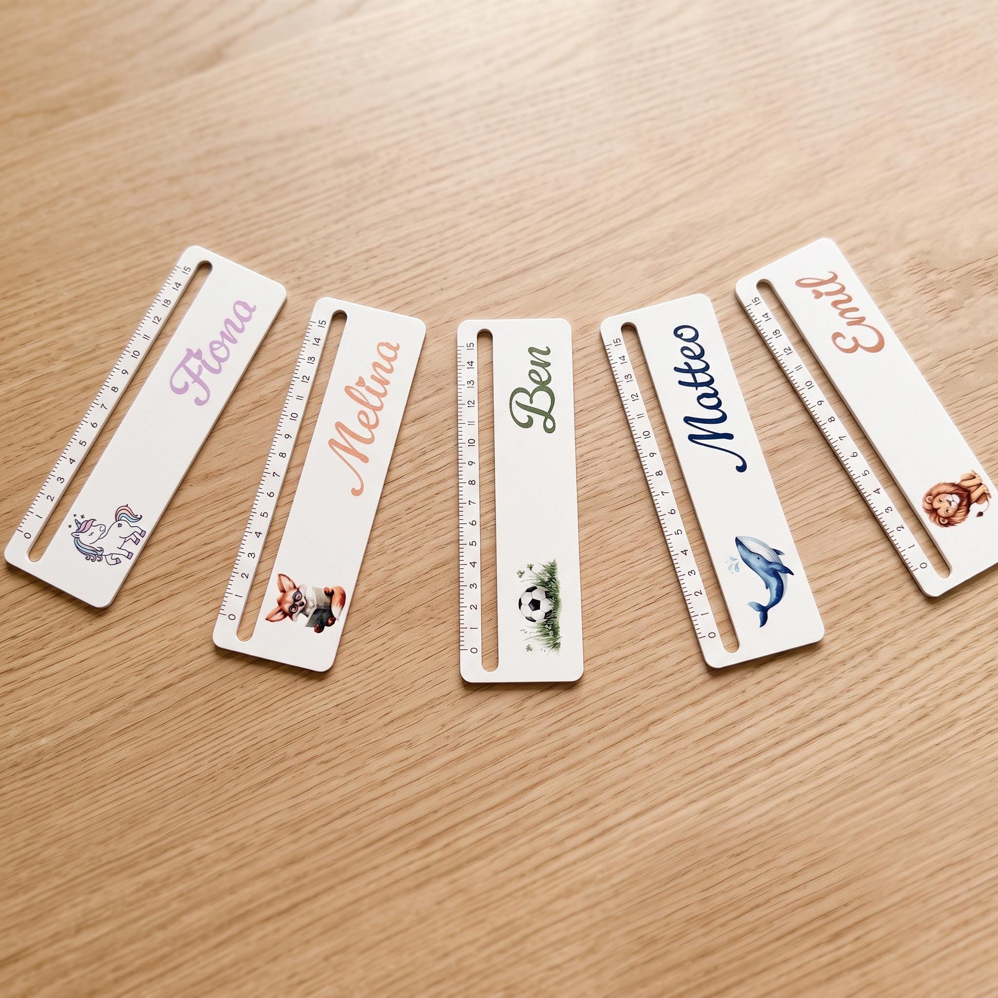 Five white rectangular rulers with black measurement markings, each personalized with a name and a different colorful illustration, laid out on a light brown wooden surface.