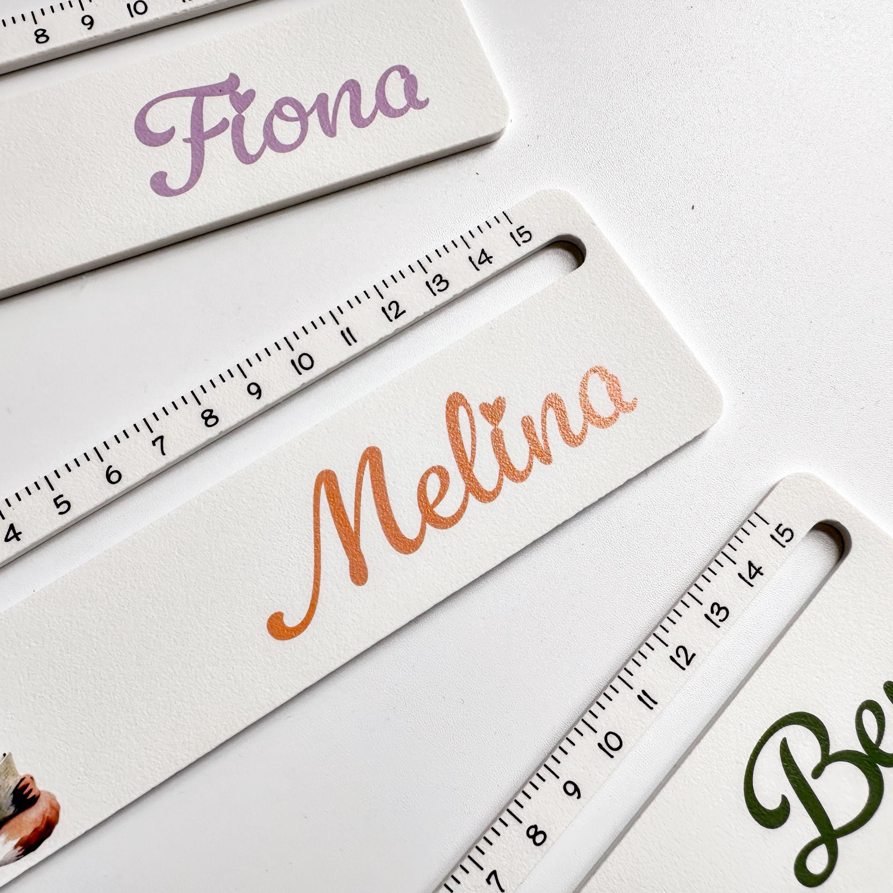 A close-up, angled view of two white rulers with black measurement markings; the top one is personalized with "Fiona" in purple, and the bottom with "Melina" in orange.