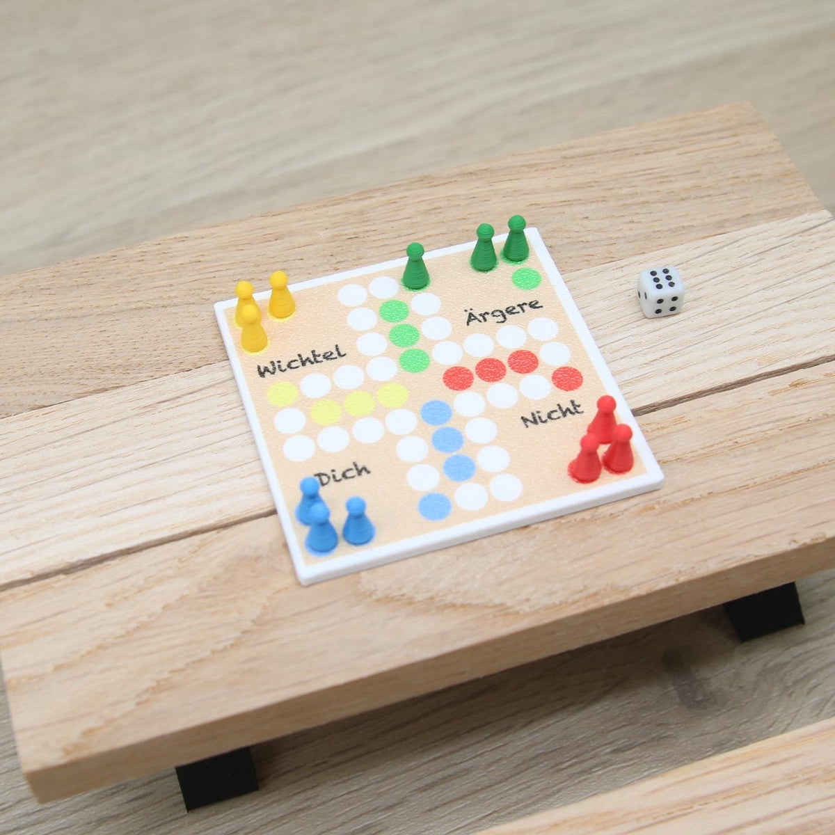 Könnte beinhalten: Ein quadratisches Brettspiel mit einer hellen, holzfarbenen Oberfläche und einem Raster aus weißen, gelben, grünen, roten und blauen Kreisen. Das Brett enthält den Text Wichtel, Ärgere, Nicht und Dich. Es umfasst gelbe, grüne, rote und blaue Spielfiguren sowie einen Würfel.