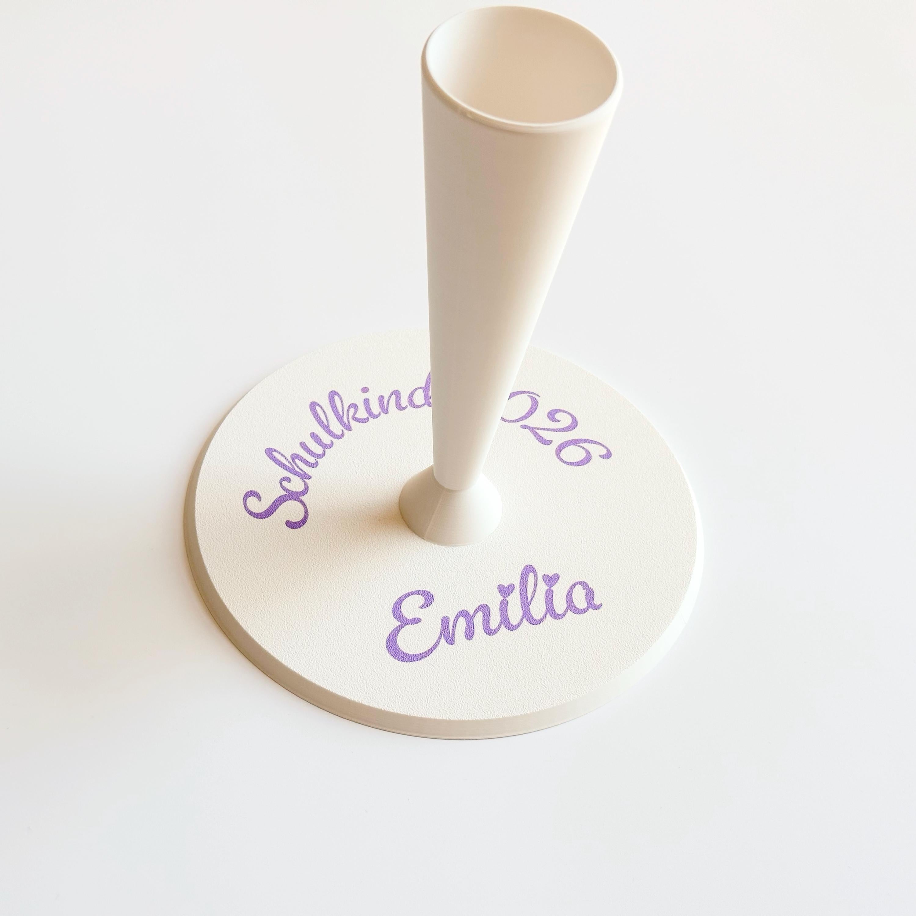 A cream-colored conical stand is inserted into a round cream-colored base, which has "Schulkind 2026" and "Emilia" printed in purple script.