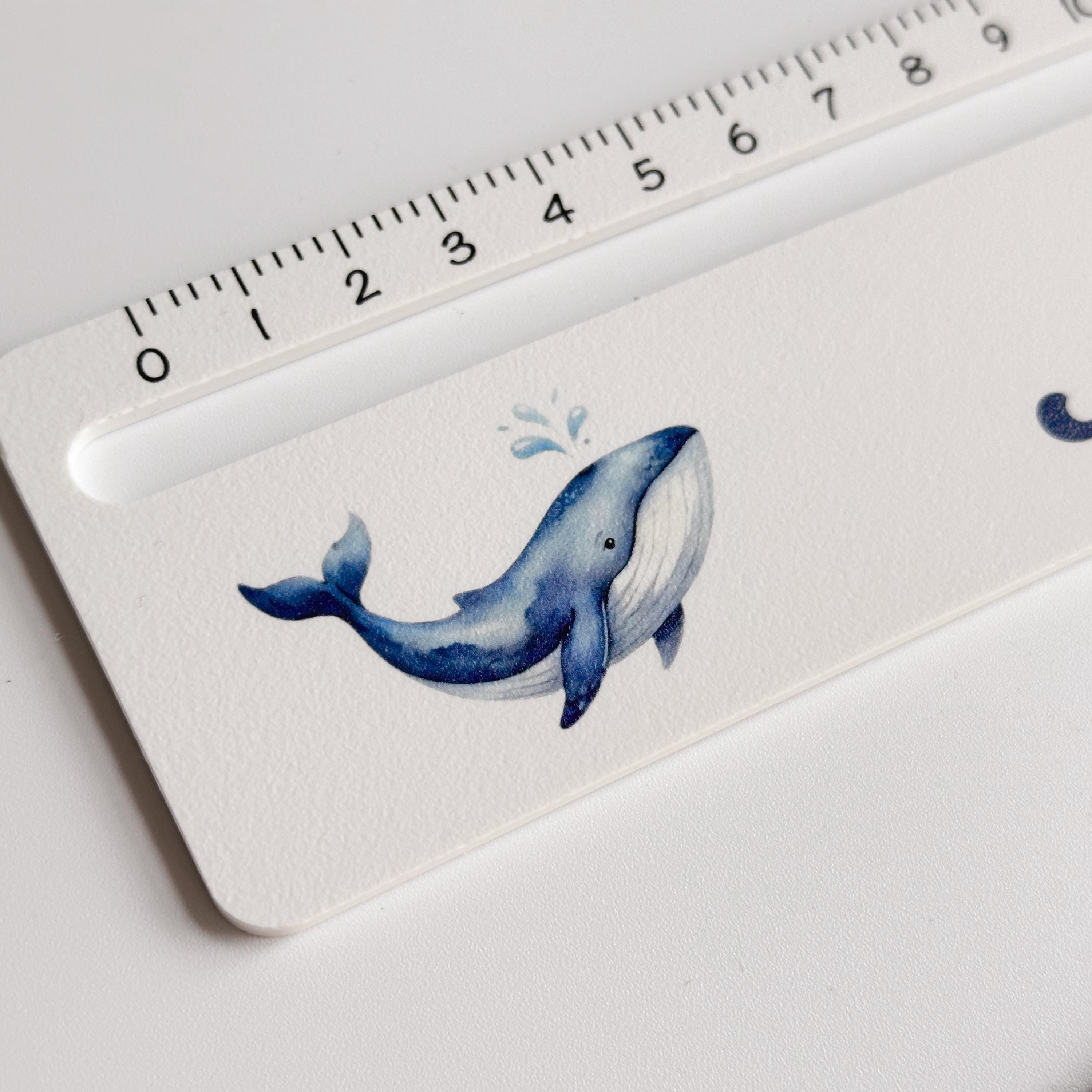 A close-up of a white ruler with black measurement markings, featuring a blue whale illustration with water splashes on the right side.