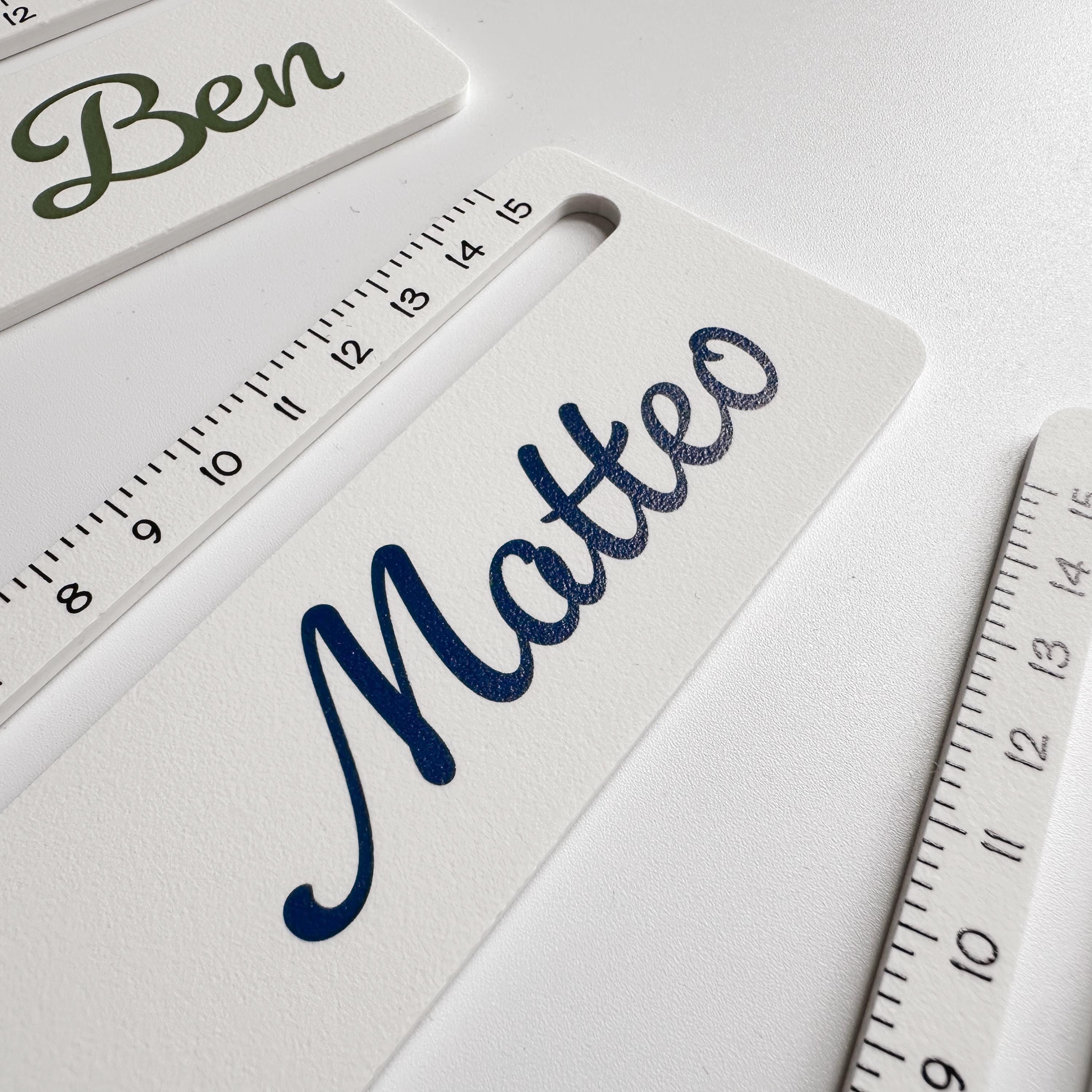 A close-up, angled view of a white ruler with black measurement markings, personalized with the name "Matteo" in dark blue script, with another ruler visible behind it.