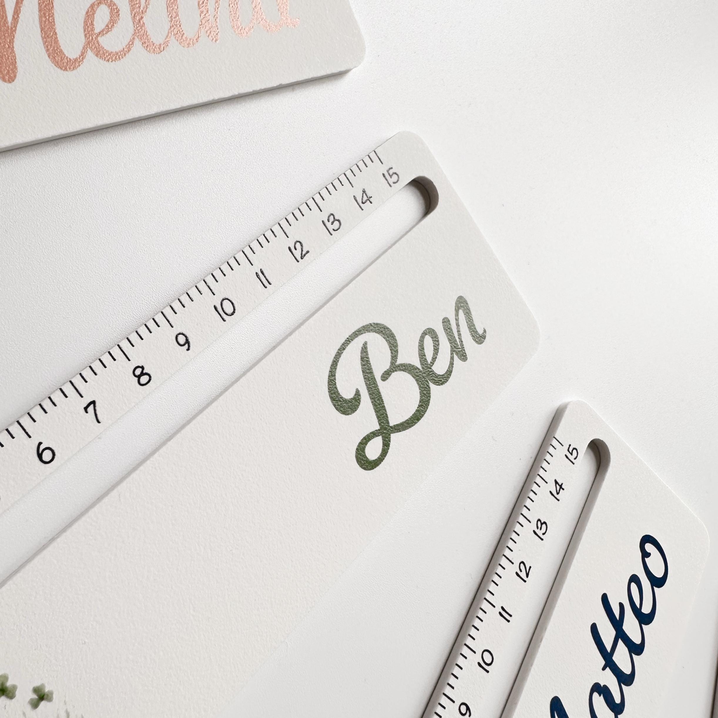 A close-up, angled view of a white ruler with black measurement markings, personalized with the name "Ben" in dark green script, with parts of two other rulers visible.
